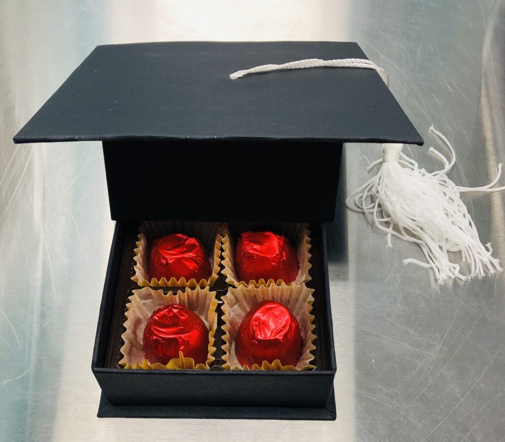 Graduation cap gift box filled with four chocolate-covered cherries wrapped in red foil, displayed on a stainless steel surface.